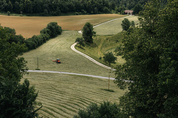 A serene scene from Radovljica with rolling green hills, freshly mowed fields, and a winding path. A tractor works the land, surrounded by dense trees and a distant house under a clear sky.