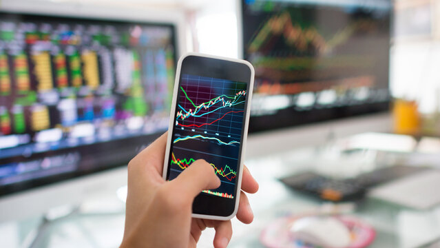 Real-time market analysis: Hand holding smartphone with financial charts, monitoring investment data on a desktop screen in a contemporary office. - Powered by Adobe
