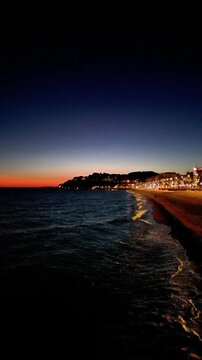 Aerial drone view of coastline town at night, moonlight reflected on sea waves, city lights, vertical background