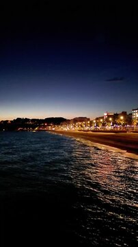 Aerial drone view of coastline town at night, moonlight reflected on sea waves, city lights, vertical background