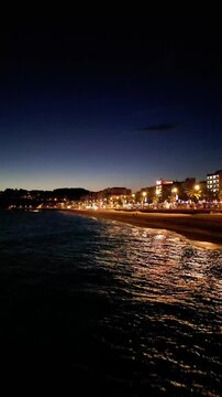 Aerial drone view of coastline town at night, moonlight reflected on sea waves, city lights, vertical background