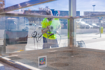 A worker in a high-visibility yellow shirt uses a power tool to remove graffiti from a glass...
