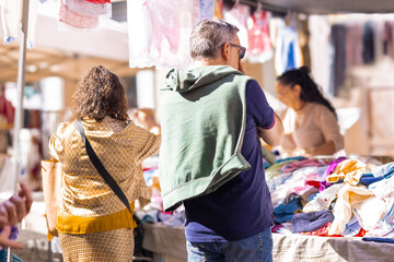 Shoppers browse through piles of colorful clothing at a bustling outdoor market in Valencia. Sunlight filters through the stalls, illuminating the vibrant textiles and the busy atmosphere of the