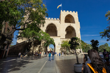 The Serranos Towers Torres de Serranos stand tall in a sunlit plaza in Valencia. People walk past the massive Gothic gateway, which features grand vaulted arches and a flag flying from its crenelated
