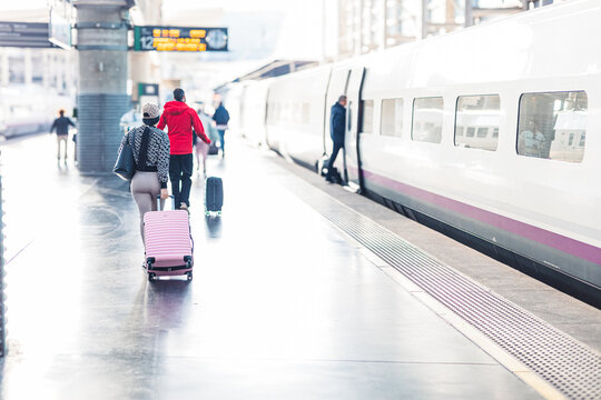 Travelers with rolling suitcases walk along a bright, modern platform toward a sleek high-speed train at Madrid Puerta de Atocha station. The white train wait for boarding under a bright, airy
