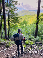 Back view of hiker with large backpack and sleeping pad stands on rocky forest trail, gazing at scenic mountain view. Lush greenery and tall trees frame distant, rolling hills under partly cloudy sky.