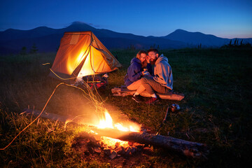 Couple hikers enjoys cozy moment by campfire, sipping hot drinks near glowing tent. Twilight sky and mountain backdrop create serene and romantic atmosphere in picturesque wilderness setting.