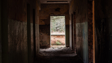 Hallway in a deserted building shows peeling walls and debris on the floor. Light comes through an open doorway at the end, revealing a view outside.
