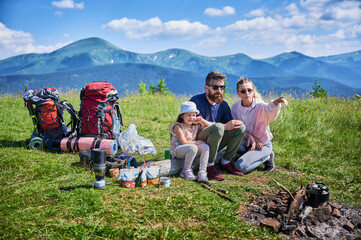 Family of three enjoys camping trip in mountains, with backpacks and camping gear around. Mother points at something in distance while father and daughter, taking in scenic view of lush green hills.
