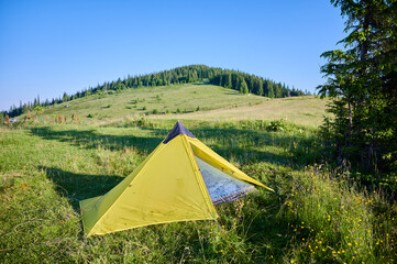 Bright yellow tent on lush grassy meadow under clear blue sky. On background gently sloping hill covered with scattered trees and shrubs, creating serene and picturesque camping scene.