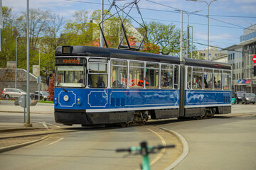 A blue and white vintage-style tram travels along a curved track in Tallinn. The tram, headed for Kadriorg, moves through an urban intersection under a bright, overcast sky.