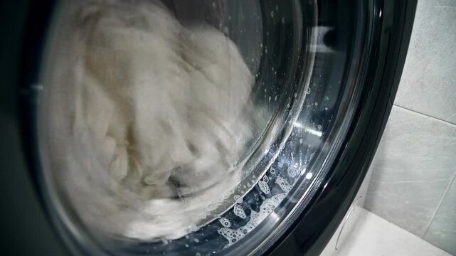 A close-up of old, yellowed gray laundry that was once white being washed in a washing machine. Bubbles of detergent drip down the glass window.