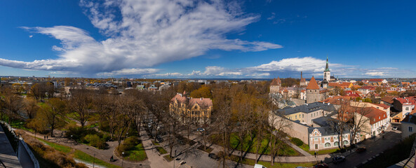 Wiide panoramic view of Tallinn, capturing the contrast between the lush parks of the lower town and the red-tiled roofs and defensive towers of the historic Old Town under a dramatic, cloud-filled
