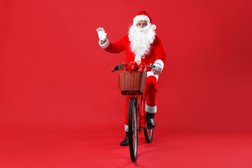 Santa Claus on bicycle with Christmas gifts in basket greeting someone against red background
