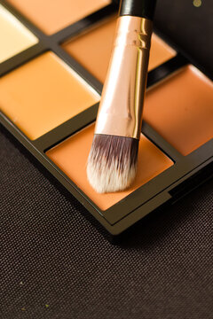 A close-up image of a professional makeup brush dipped into a multi-shade cream foundation or concealer palette. 
