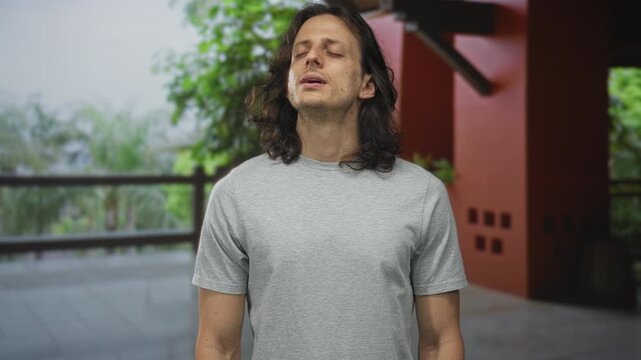 Man with long hair and eyes closed, bare face turned upward on a building terrace, breathing slowly and standing still; serenity meditation.