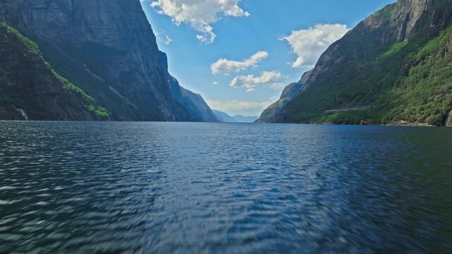 Aerial view flying low above Lysefjord near Lysebotn, Norway. Calm blue fjord water is framed by steep granite cliffs and lush green mountains on a bright summer day.