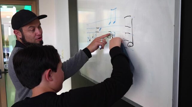Student practices rhythm writing on the board while the teacher offers guiding and corrections in a playful music class. Music notation, solfege study, structured rhythm education