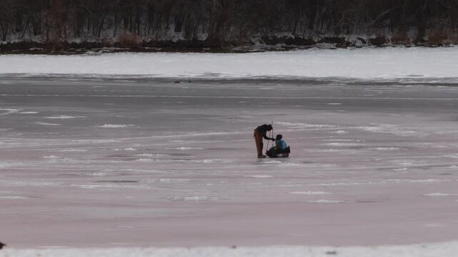 An unrecognizable person far in the distance walking over frozen lake water and pulling his child sitting on a sled on the ice as viewed through a long zoom lens.
