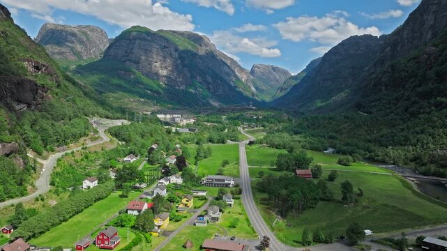 Drone flying above the green valley of Lysebotn near Lysefjord, Norway. The landscape features farmland, village roads and towering mountains in summer light.