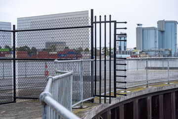 Security fencing and restricted access at the waterfront in Docklands, Melbourne, Australia, with industrial silos, port infrastructure, and modern buildings visible across the water of Yarra River.