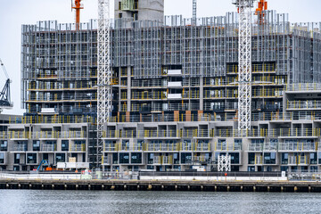 A large multi-storey building under construction along a waterfront, wrapped in scaffolding and safety screens, with tower cranes rising above the structure. High-density residential construction