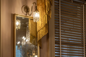 An elegant vintage interior detail featuring an ornate wall sconce with a crystal glass shade, reflected in a gold-framed mirror. Heavy gold tasselled drapery. A warm, luxurious atmosphere