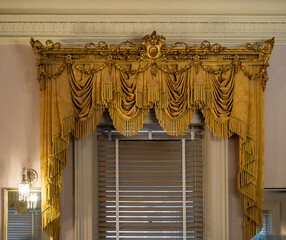 An ornate gold-colored window valance with elaborate drapery, tassels, and carved decorative details, framing a traditional interior window with blinds behind. Classical interior design, historic luxu