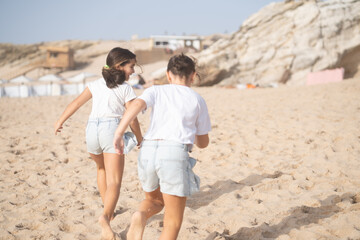Rear view of cheerful girls in casuals running on sandy beach enjoying summer vacation with mountains in background during sunny day