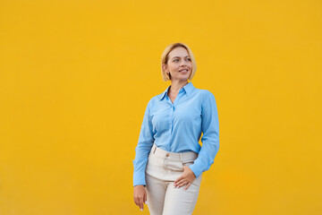 Confident blond businesswoman in stylish blue shirt standing against yellow background smiling while looking away with professional and modern appearance