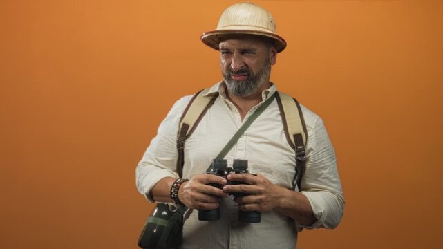 Man holding binoculars, wearing a pith helmet and backpack in orange studio; curiosity travel adventure.