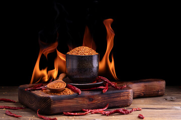 Chili powder in black bowl and wooden spoon with garlic, shallots, garden spicy spice mix. Asian food ingredients arranged on wooden table. Flame background, spicy food concept.