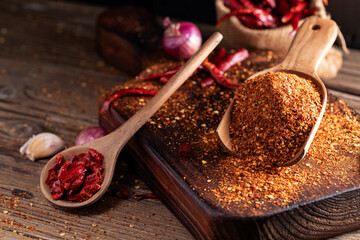 Chili powder in a wooden bowl and with a wooden spoon, along with garlic, shallots, and a mix of Asian spices, are arranged on a wooden table, conveying the concept of spicy food.