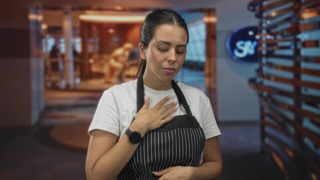 Young brunette woman in striped apron brings hand to mouth over a tasting sample while standing by a hotel building lobby display; quiet relief.
