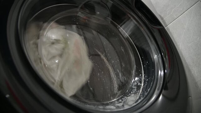 A close-up of old, yellowed gray laundry that was once white being washed in a washing machine. Bubbles of detergent drip down the glass window.