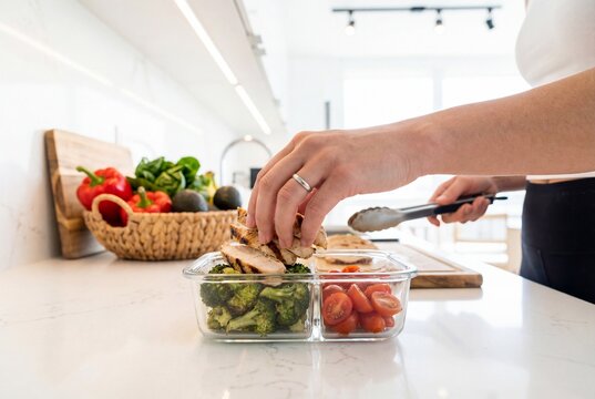 Woman packing grilled chicken and vegetables into glass container for healthy meal prep lunch