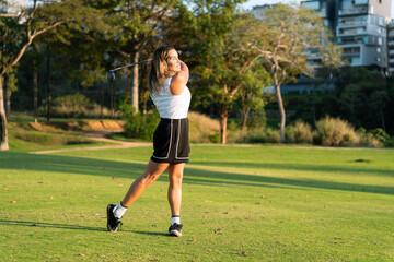 Obraz premium A latin athletic woman performs a powerful golf swing on a lush green course under golden hour sunlight