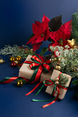 Christmas decor with handmade gift boxes, red poinsettia, decorative fir branch, bells and balls for tree decoraton on blue vertical background. New Year winter holiday composition with presents.