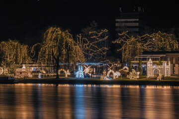 A beautifully lit riverside park filled with festive light installations, creating a warm and enchanting night scene reflected on the water.