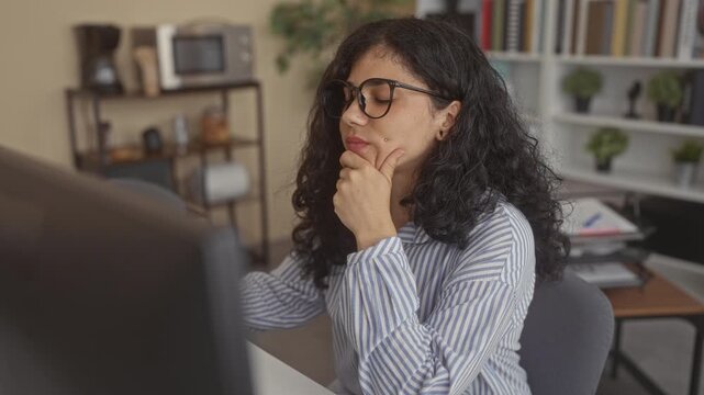 Woman resting hand on chin at office building desk, looking at computer screen while wearing glasses and a striped shirt; thoughtful pondering.