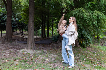 Fototapeta premium Young woman holding child while pointing at tree in a park