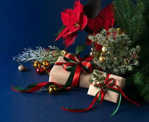 Christmas decor with handmade gift boxes with bow, red poinsettia, decorative fir branch, bells and balls for tree decoraton on blue background. New Year winter holiday composition with presents.