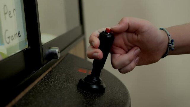 Close-up of a hand gripping and moving an arcade joystick while pressing the red top button on a game cabinet, showing classic controller interaction and indoor arcade gameplay.