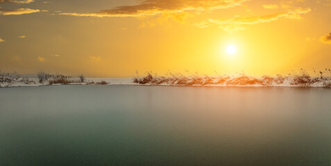 Beautiful landscape of a frozen lake covered with snow and reeds under a golden sunset sky in winter