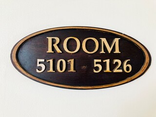 oval wooden room directory sign with gold lettering on a white wall