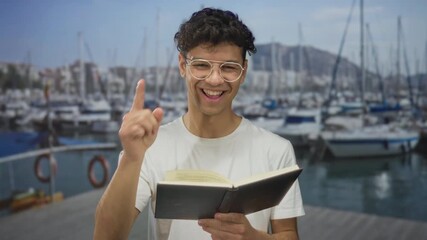 Young hispanic man wearing glasses holds an open book and points finger to text while reading on a wooden port dock; curiosity.