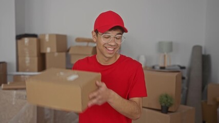 Man holding cardboard box and smiling in building wearing red cap and glasses amid stacked moving boxes; joy.