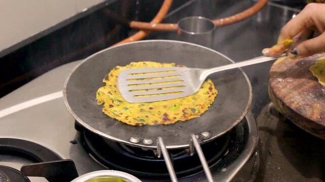 Masala roti cooking in Indian home kitchen with woman flipping spiced flatbread on hot tawa over gas stove, traditional vegetarian food preparation showing daily household routine.