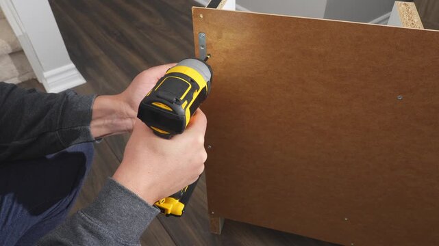 Individual is using a power drill to secure a metal bracket to a wooden surface, demonstrating assembly techniques in a well-lit indoor space