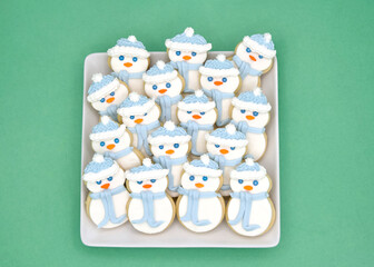 Top above view of square porcelain plate filled with homemade sugar cookies snowmen with marshmallow fondant and royal icing. On a light green background.